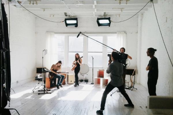Understanding The Small Business Landscape in Melbourne 3 A music band is performing in a bright, white studio with large windows. A person is operating a boom microphone while another films with a camera. The band members, playing instruments including guitar and drums, create an electrifying atmosphere as the scene captures the essence of a small business in Melbourne. | Ven Agency