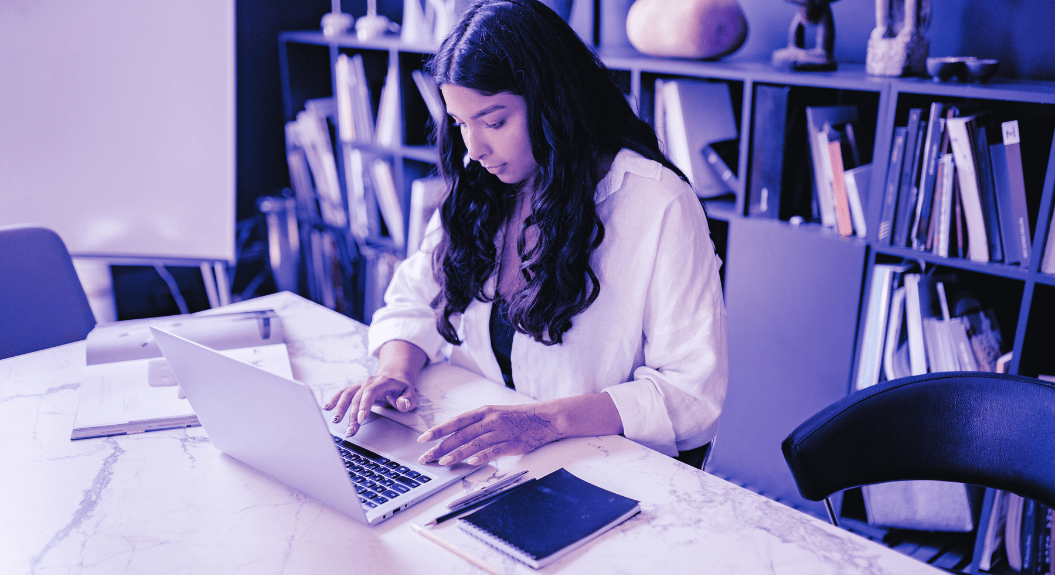 The Ultimate Guide to Website Layout and UX Design 3 A woman with long hair focuses on UX design at a marble table in an office space. The background features a large bookshelf filled with various items. A notepad and pen rest on the table, complementing her work on crafting the ultimate guide to website layout. The image is tinted purple. | Ven Agency