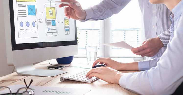 Choosing the Right Web Design Agency and Development in Melbourne 6 Two people collaborate on a web development project for a design agency. In the naturally lit Melbourne office, one points at wireframe designs on the monitor while the other types. Documents and a glass of water are scattered across the desk, as sunlight streams through the windows. | Ven Agency