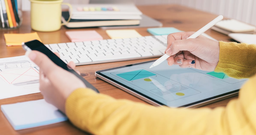 Choosing the Right Web Design Agency and Development in Melbourne 8 A person in a yellow sweater uses a stylus to design on a tablet displaying a web development wireframe. A smartphone is held in the other hand. On the wooden desk of this Melbourne web design agency, you'll find a keyboard, sticky notes, and a cup with pens. | Ven Agency