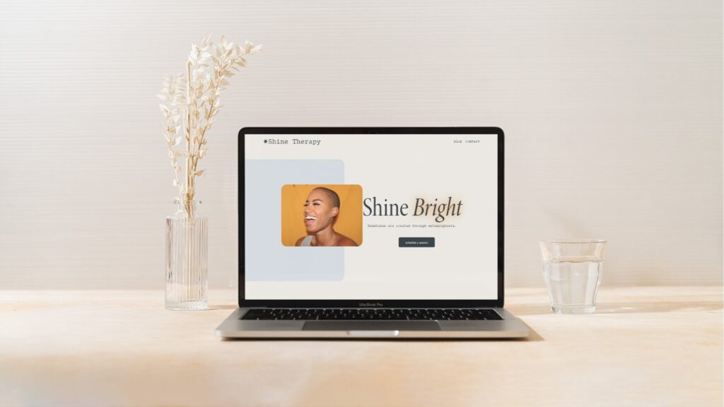 Is Squarespace the Right Choice for Business Website? 2 A laptop on a wooden table showcases a business website with the text "Shine Bright" beside an image of a smiling person. A tall vase with dried flowers sits to the left, and a glass of water to the right. This elegant setup, perhaps crafted on Squarespace, is framed by a neutral-toned background. | Ven Agency